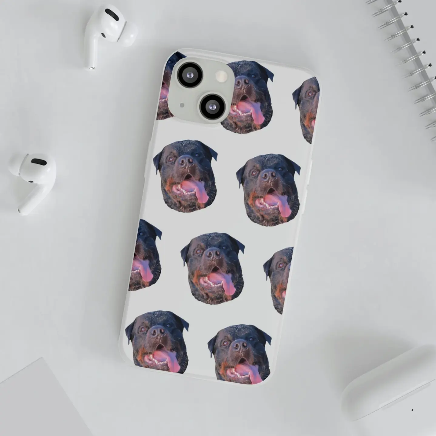 Overhead view of a white custom phone case patterned with Rottweiler faces, accompanied by wireless earbuds and a notebook